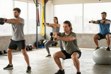 A vibrant gym environment showcasing the benefits of functional training. In the foreground, a diverse group of three individuals engaged in exercises, wearing modest athletic wear, such as a fitted t-shirt and shorts. One person is lifting a kettlebell, another is performing a squat with dumbbells, and the third is demonstrating a balance exercise on a stability ball. The middle ground features exercise equipment such as resistance bands and free weights, highlighting versatility. The background includes large windows allowing natural light to flood the space, creating a warm and energetic atmosphere. The setting appears motivating and lively, emphasizing health and vitality. Use soft, bright lighting with a slightly blurred focus on the background to enhance the subjects in action, reflecting a sense of community and wellness.